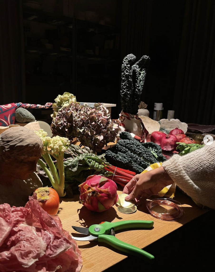 Hands arranging fruit, vegetables and flowers on a long table for an event table setting by Elena Pelosi.