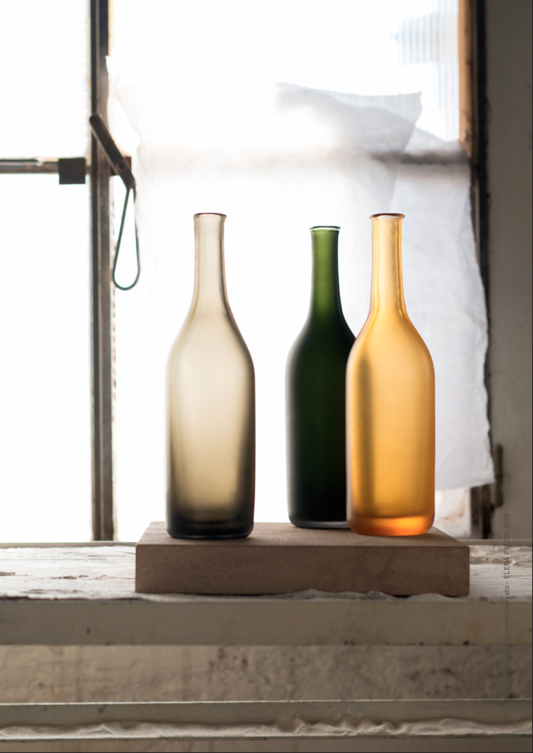 Three Venini glass bottles in smoky grey, green and amber on a wooden block by a window, Alla Morandi collection.