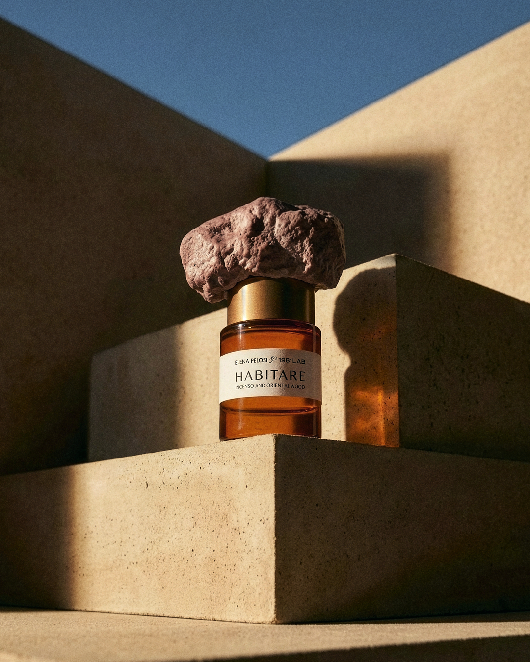 HABITARE fragrance bottle topped with a pink stone, on sunlit concrete blocks.