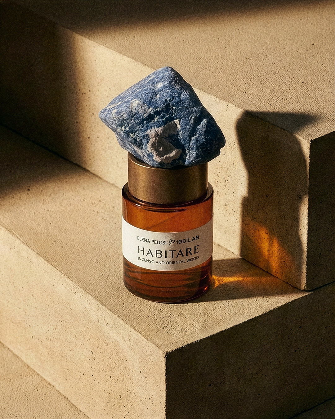 HABITARE fragrance bottle topped with a blue mineral, on a concrete step in raking light.