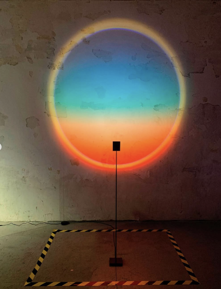 Circular halo of coloured light projected on a raw wall above a slim floor lamp, Halo Project at Lampo Milano.