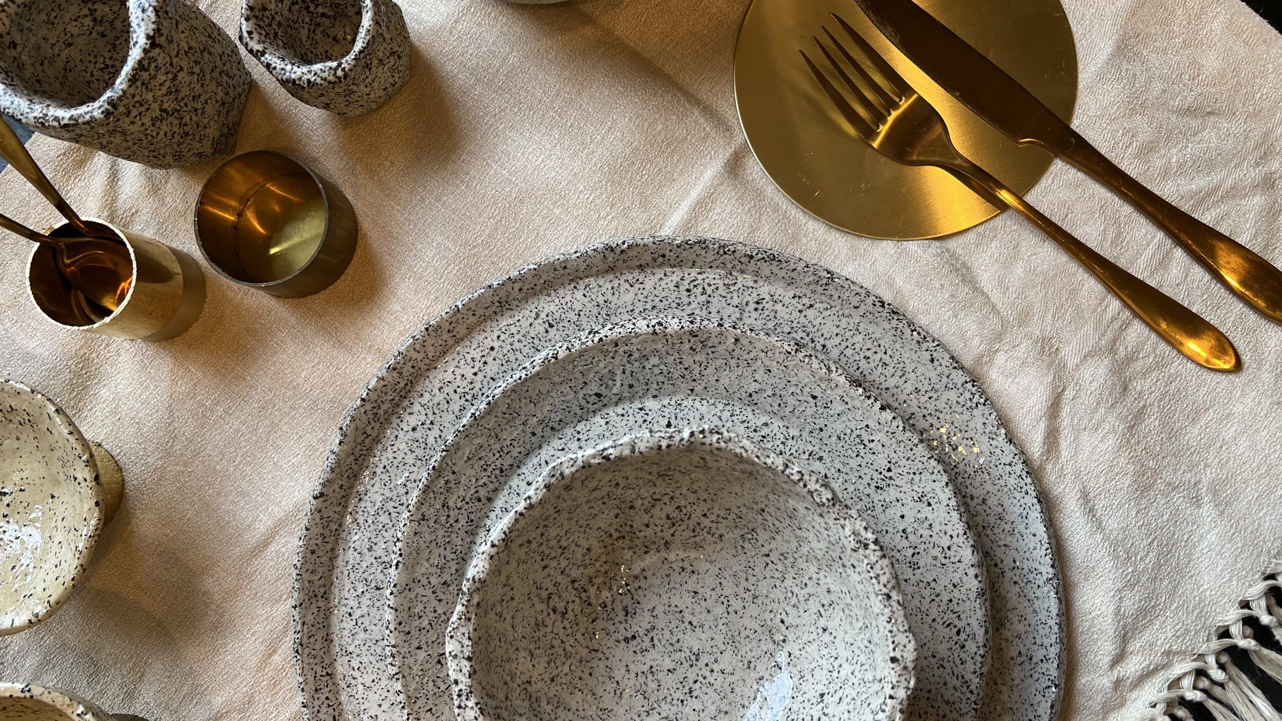 MINERALIA speckled ceramic plates and bowls with gold cutlery on a linen tablecloth.