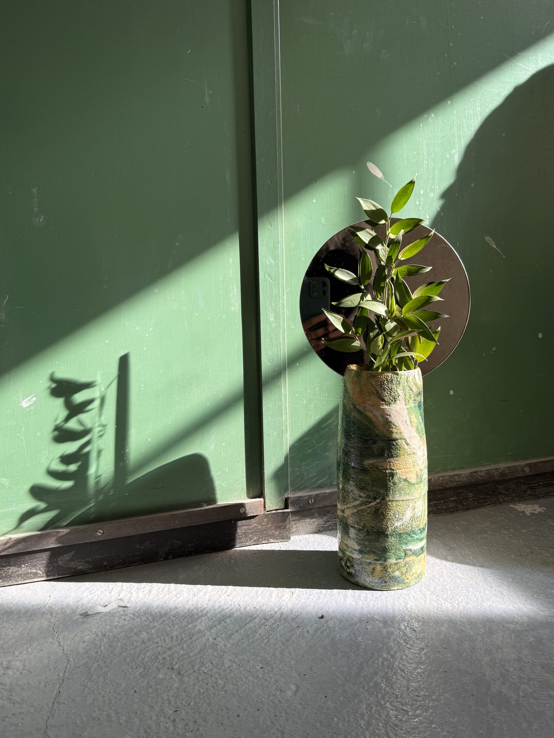 Panta Rei marbled ceramic vase with a leafy branch and a brass disc, against a green wall.
